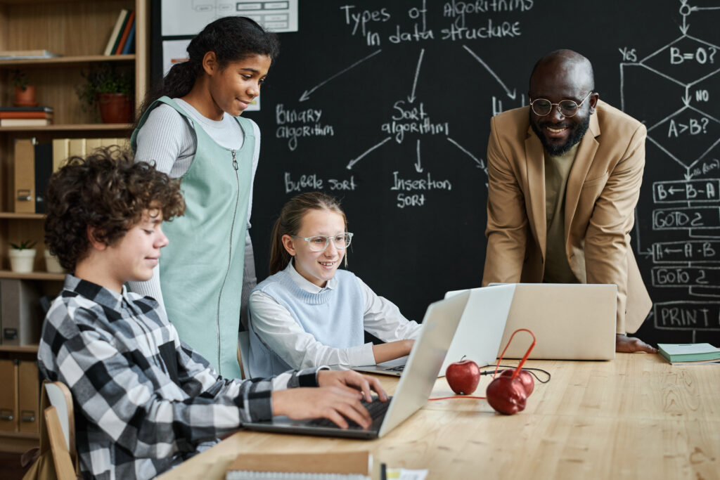 Group of children learning to work with computer program using laptop computers at table together with teacher at lesson in a flipped classroom model