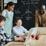 Group of children learning to work with computer program using laptop computers at table together with teacher at lesson in a flipped classroom model