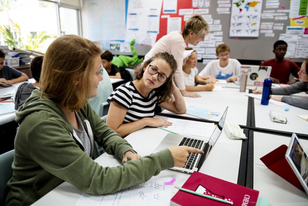 Group of student sitting at desks and working together at computers while a teacher stands in the classroom, showing the concept of collaborative learning.
