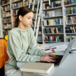 Teenage female student studying on her laptop while sitting in a library, showing the concept of schools adopting blended learning strategies.