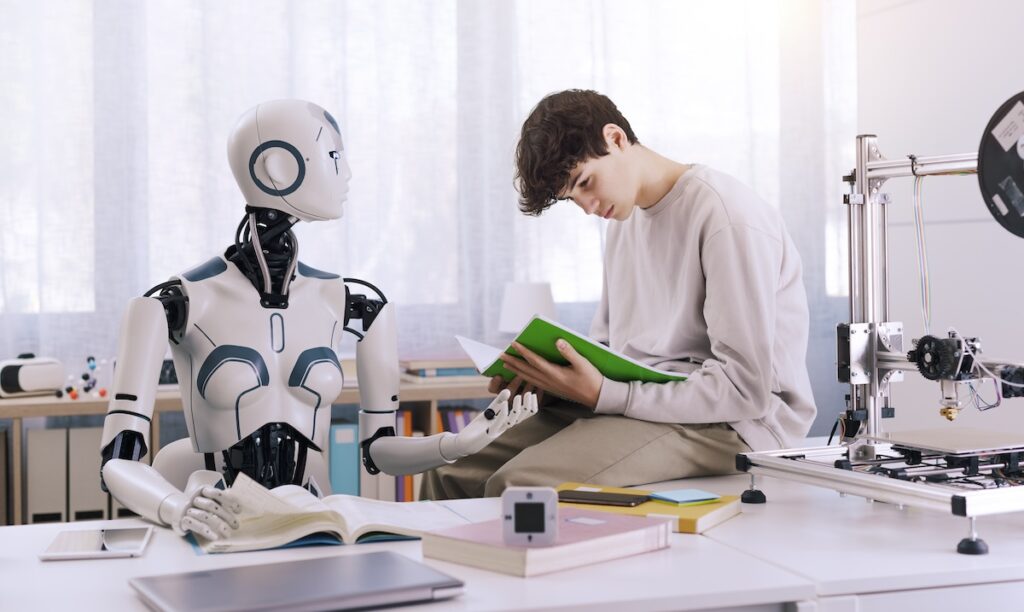 Male student sitting on desk reading a book with a robot teacher standing next to him demonstrating the concept of AI in schools.