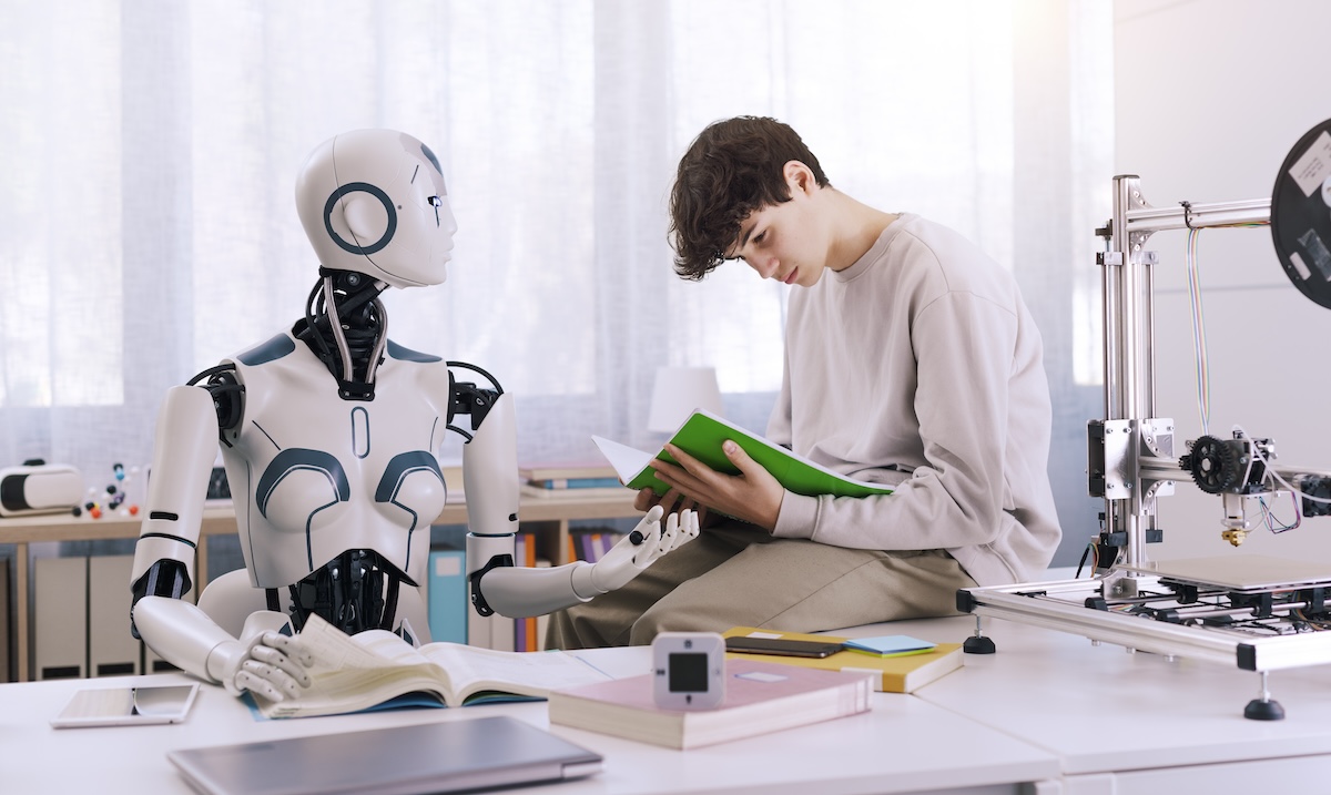 Male student sitting on desk reading a book with a robot teacher standing next to him demonstrating the concept of AI in schools.