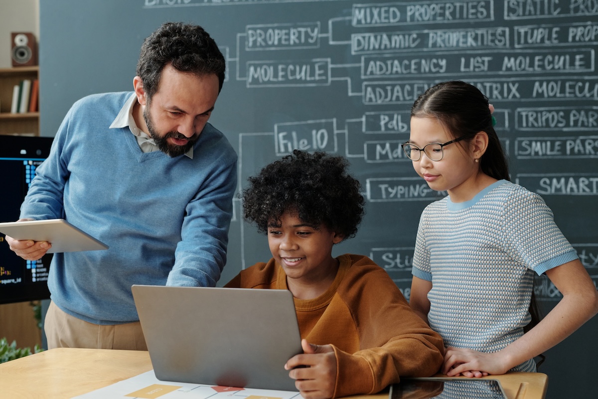 Teacher offering guidance to two focused students working on laptop during class. Blackboard with scientific terms visible in background showcasing an educational environment and highlighting the concept of how to use AI in the classroom.