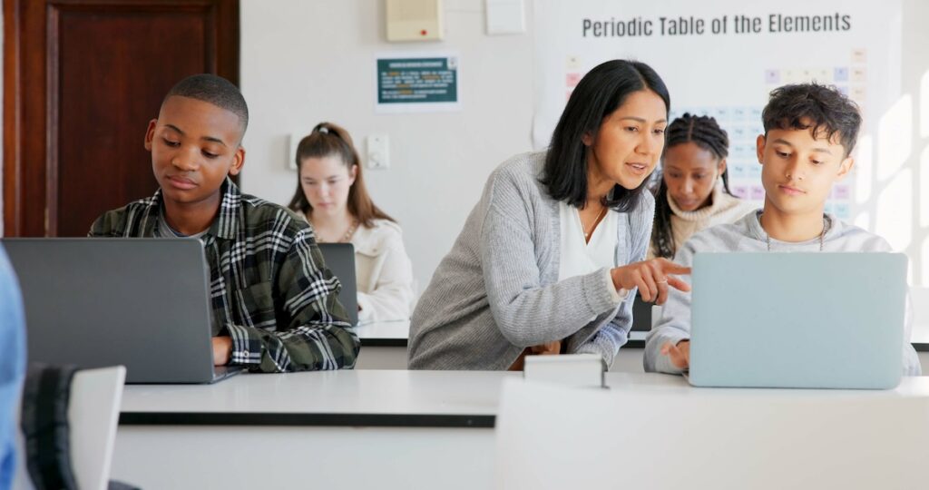 Four teen students sitting in a classroom using their laptops while a female teacher leans over a student to point at something on their computer screen, demonstrating the concept of customized and personalized learning.