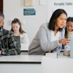 Four teen students sitting in a classroom using their laptops while a female teacher leans over a student to point at something on their computer screen, demonstrating the concept of customized and personalized learning.