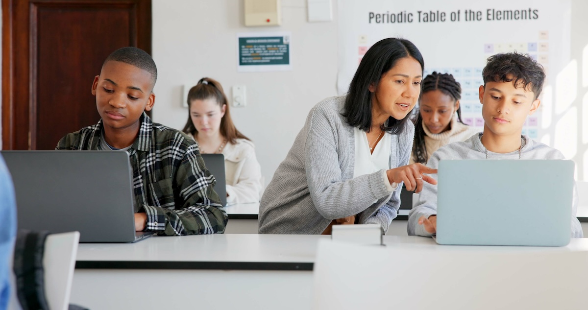 Four teen students sitting in a classroom using their laptops while a female teacher leans over a student to point at something on their computer screen, demonstrating the concept of customized and personalized learning.