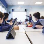 Children sitting at tables in the classroom with their tablets while turning to face their teacher at the board, showing the concept of how technology helps students learn.