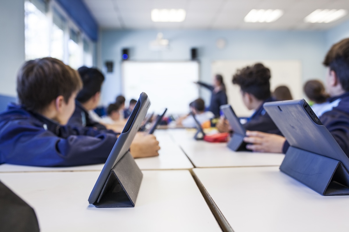 Children sitting at tables in the classroom with their tablets while turning to face their teacher at the board, showing the concept of how technology helps students learn.