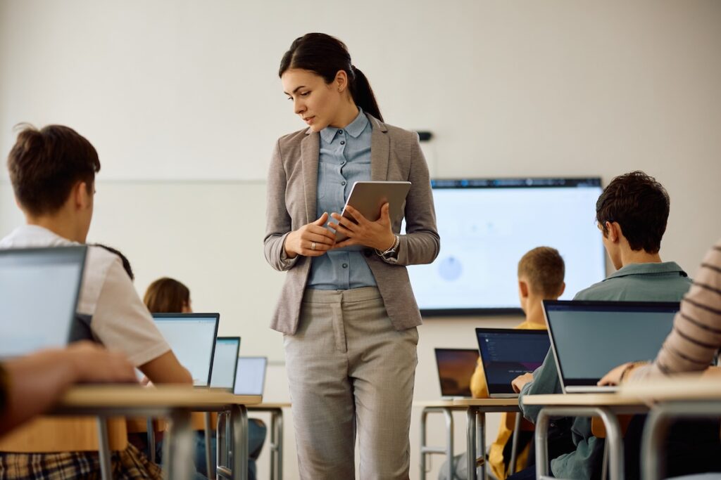 Female teacher standing in classroom in between rows of student desks, holding a tablet, while high school students sit at their desks in front of their laptops, facing the front of the class. Showing the concept of how to prevent AI misuse in education.