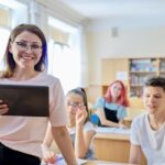 Smiling female teacher standing in front of a classroom facing the camera while holding a tablet as students sit at desks in the background