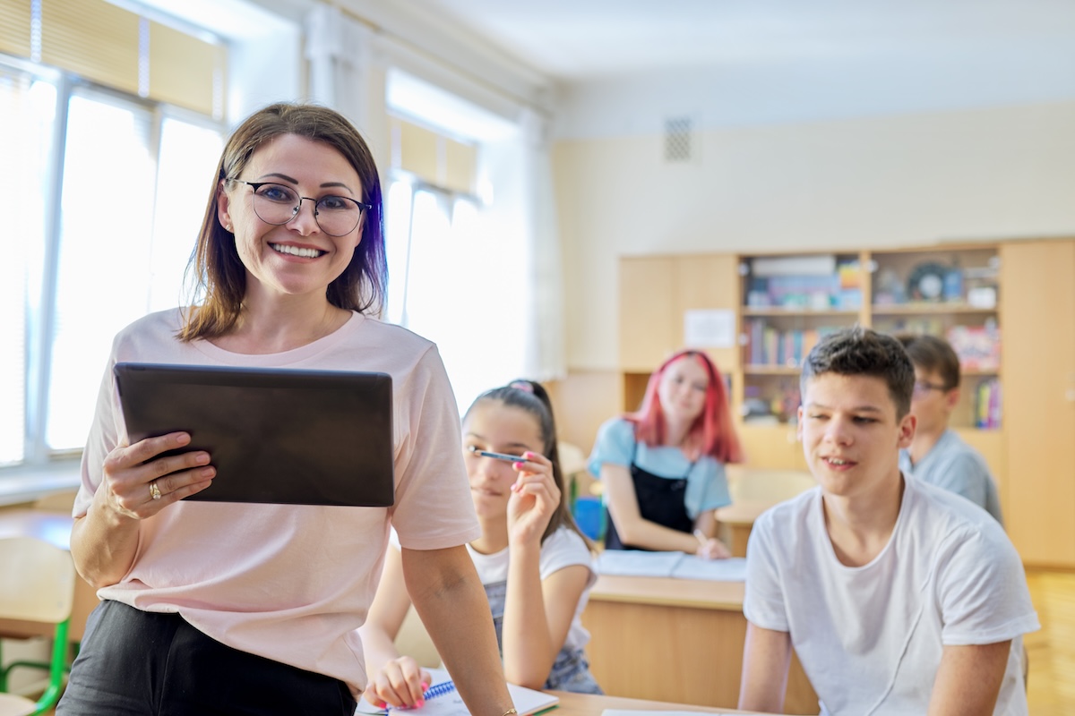 Smiling female teacher standing in front of a classroom facing the camera while holding a tablet as students sit at desks in the background