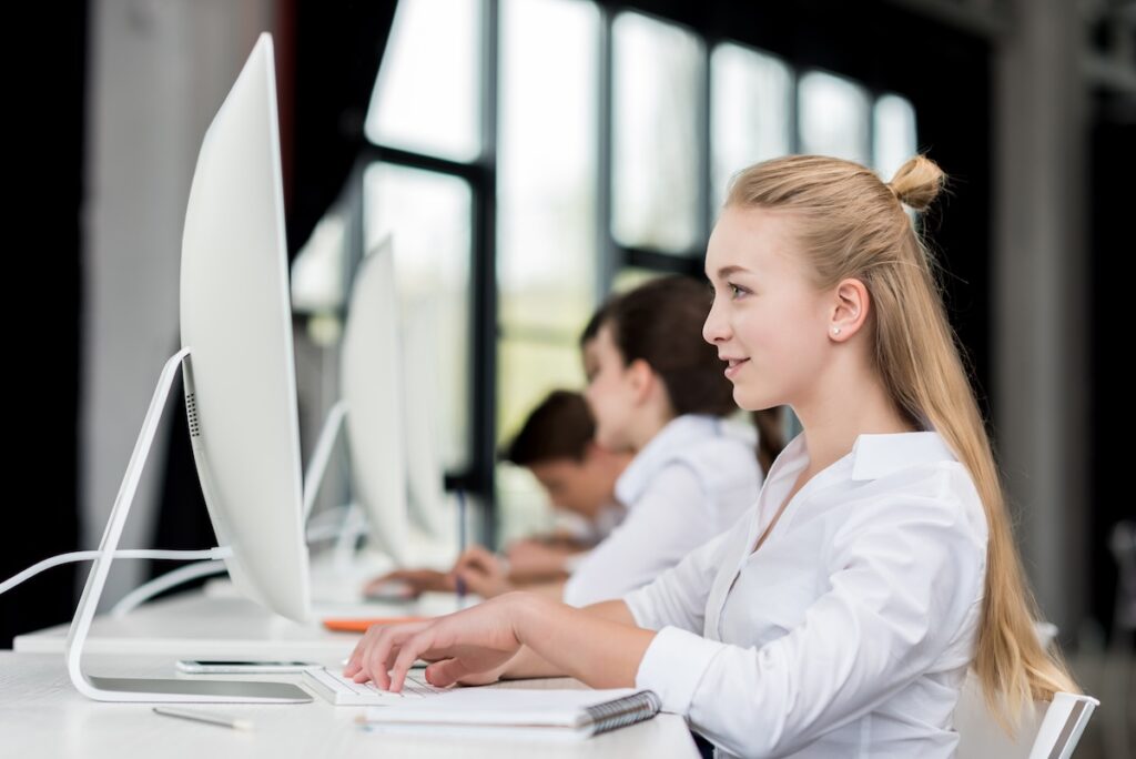 side view of smiling teen girl sitting and her classmates sitting in a row while taking an exam on a computer, showing the concept of how to reduce students' test anxiety.