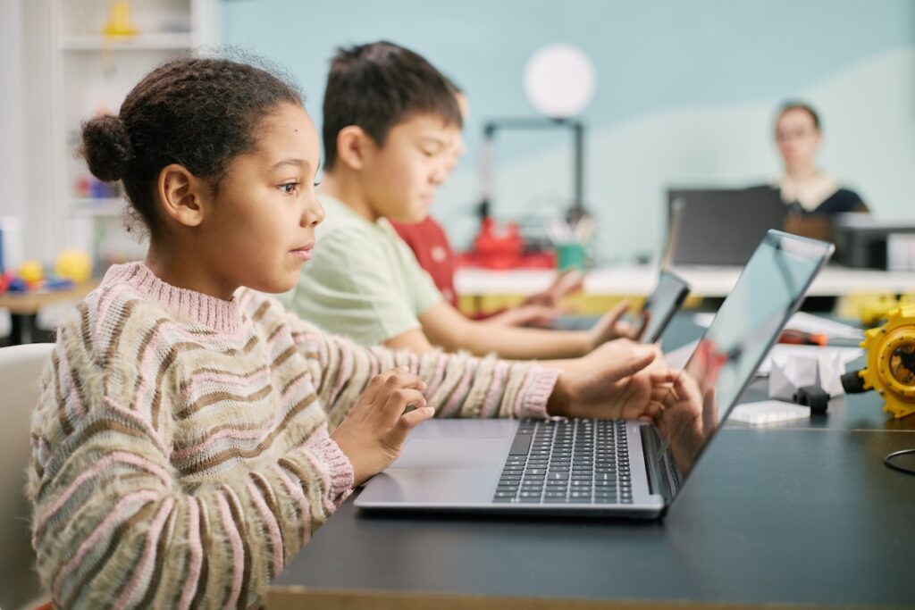 Two young students, one male and one female, sitting at a table in a classroom using their laptops, demonstrating the concept of utilizing open source assessment software. Out of focus in the background there is a teacher sitting at their desk.