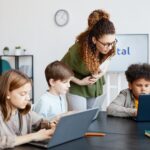 Three young students are sitting at a desk using their laptops, showcasing the concept of building equity in education. A female teacher stands behind and leans over one students computer to check their progress.