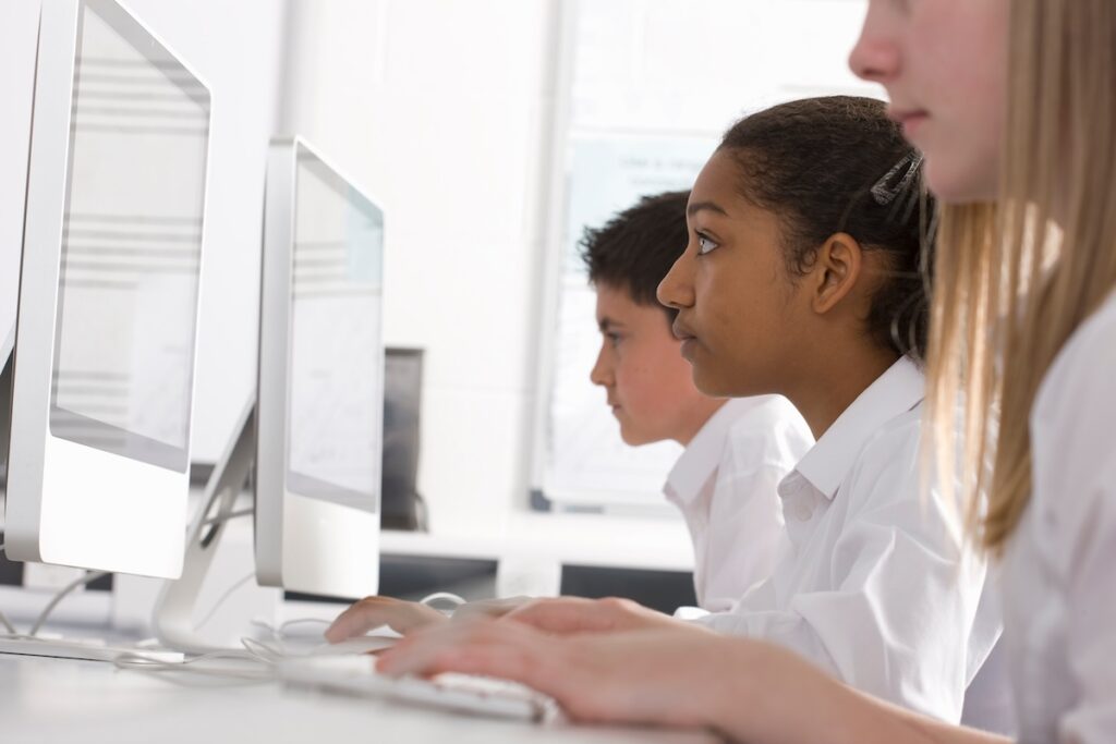 Side view of two female students and one male student sitting in a row in a computer class while looking at their desktop computers, showing the use of open educational resources in a school context.