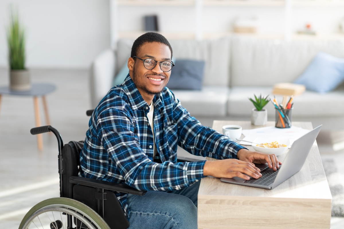 Smiling man wearing glasses sits in his wheelchair at a desk with his hands on a laptop while taking an exam designed for digital accessibility.