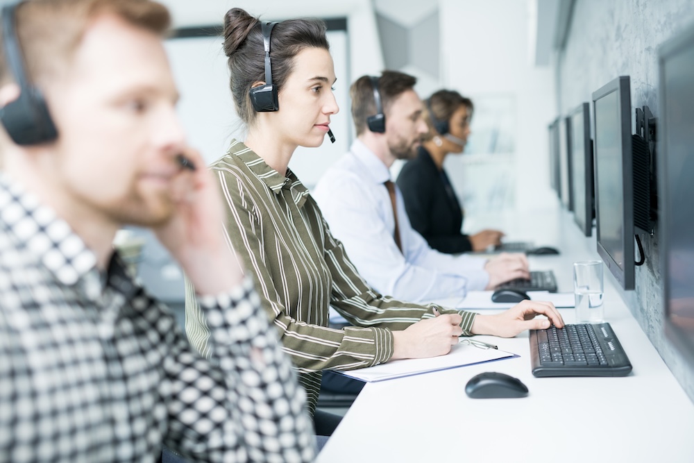 Seitenansicht mehrerer Helpdesk-Mitarbeiter, die mit Headsets in einer Reihe sitzen, auf Computerbildschirme blicken und Headsets tragen, um das Konzept der Live-Online-Aufsicht zu veranschaulichen