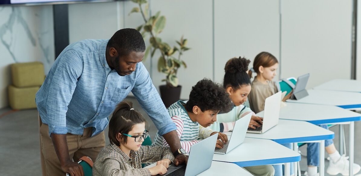 Ein Lehrer steht in einem Klassenzimmer hinter einer Reihe von Schülern. Die Schüler arbeiten an ihren Laptops, während sich der Lehrer vorbeugt, um dem ersten Schüler in der Reihe zu helfen.