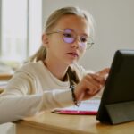 Une jeune fille à lunettes, assise à un pupitre en bois, utilise une tablette dans une salle de classe, entourée de ses camarades