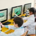 Grupo de escolares contemporáneos sentados frente a pantallas de ordenador y participando en una clase de ciencias basada en un juego.