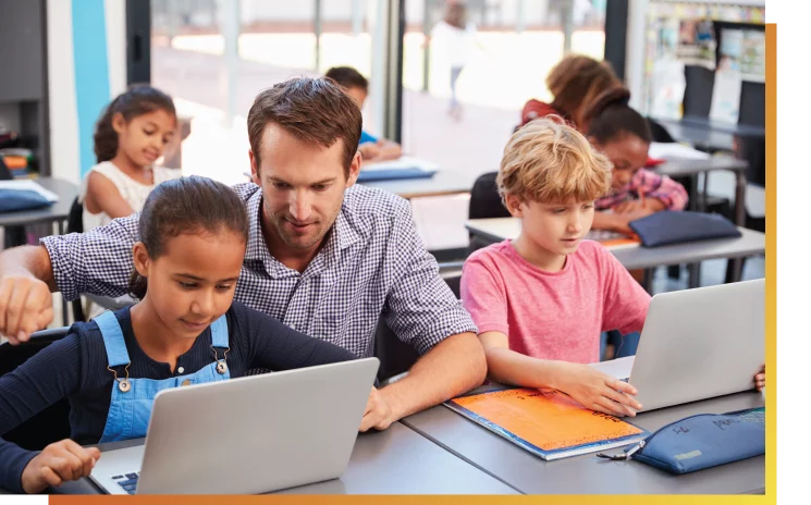Ein männlicher Lehrer hilft einer jungen Schülerin bei der Verwendung webbasierter Software auf einem Laptop, während andere Kinder im Hintergrund Geräte verwenden.