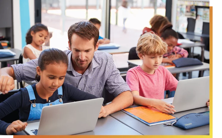 Male teacher helping a young female student using web-based software on a laptop with other children using devices in the background.