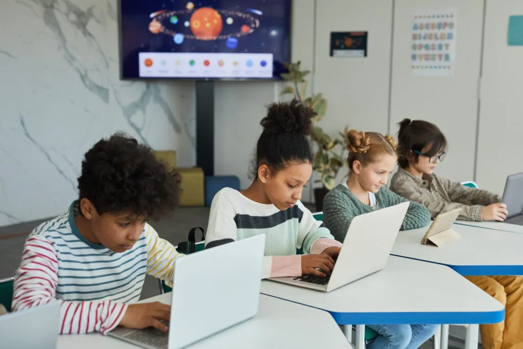 Diverse group of children sitting in row at school classroom and using laptops and computer adaptive assessment tools.