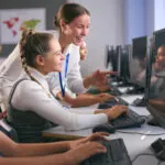 Group Of Secondary Or High School Students At Computers In IT Class With Female Teacher demonstrating the concept of adaptive learning