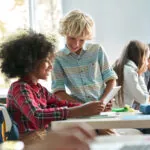 Happy smiling Caucasian boy and African American girl schoolchildren studying together using tablet device in classroom. Groups of schoolchildren working on task. Using technologies for education showing the concept of digital education access
