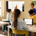 Vista posteriore di una ragazza seduta in un'aula scolastica davanti a un computer portatile con un'equazione matematica sullo schermo, che alza la mano mentre l'insegnante spiega alla classe il concetto di valutazione diagnostica in matematica.