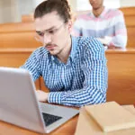 College age male student sitting in a lecture hall taking a summative evaluation on his laptop.