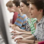 Vista di profilo di quattro bambini seduti in fila ai banchi mentre digitano sui computer della scuola durante lo svolgimento di test adattivi.