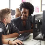 Una insegnante e un giovane studente siedono uno accanto all'altra a una scrivania davanti a un computer e si guardano sorridendo. Mostra il concetto di come le valutazioni digitali supportino gli studenti SEND.