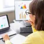 Woman wearing glasses sitting at a desk looking at a laptop and desktop monitor that each show computer-based reports on the screen.