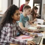 Students sitting in a horizontal row at a classroom table while working at computers, demonstrating the concept of CAT testing