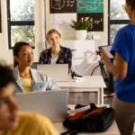 High school students sitting at their desks in from of their laptop in a classroom with a female teacher standing in the middle of the room holding a tablet, demonstrating how teachers check for AI in student work