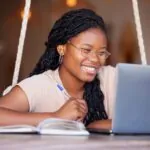 Smiling female college student sitting in front of a laptop while holding a pen and taking notes in a notebook