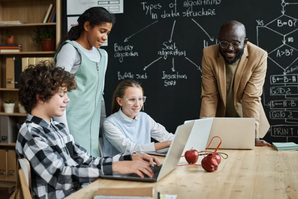 Group of children learning to work with computer program using laptop computers at table together with teacher at lesson in a flipped classroom model