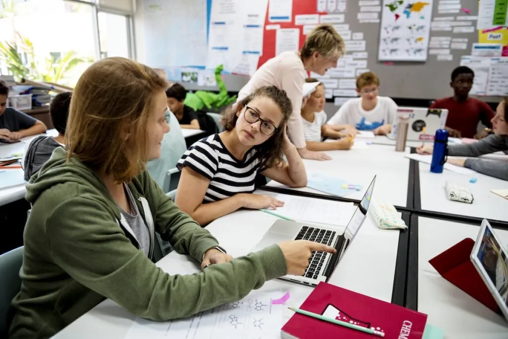 Group of student sitting at desks and working together at computers while a teacher stands in the classroom, showing the concept of collaborative learning.