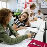 Group of student sitting at desks and working together at computers while a teacher stands in the classroom, showing the concept of collaborative learning.