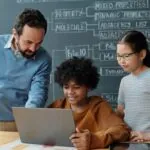 Teacher offering guidance to two focused students working on laptop during class. Blackboard with scientific terms visible in background showcasing an educational environment and highlighting the concept of how to use AI in the classroom.