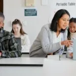 Four teen students sitting in a classroom using their laptops while a female teacher leans over a student to point at something on their computer screen, demonstrating the concept of customized and personalized learning.