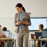Female teacher standing in classroom in between rows of student desks, holding a tablet, while high school students sit at their desks in front of their laptops, facing the front of the class. Showing the concept of how to prevent AI misuse in education.