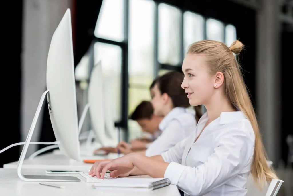 side view of smiling teen girl sitting and her classmates sitting in a row while taking an exam on a computer, showing the concept of how to reduce students' test anxiety.