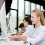 side view of smiling teen girl sitting and her classmates sitting in a row while taking an exam on a computer, showing the concept of how to reduce students' test anxiety.
