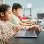 Two young students, one male and one female, sitting at a table in a classroom using their laptops, demonstrating the concept of utilizing open source assessment software. Out of focus in the background there is a teacher sitting at their desk.
