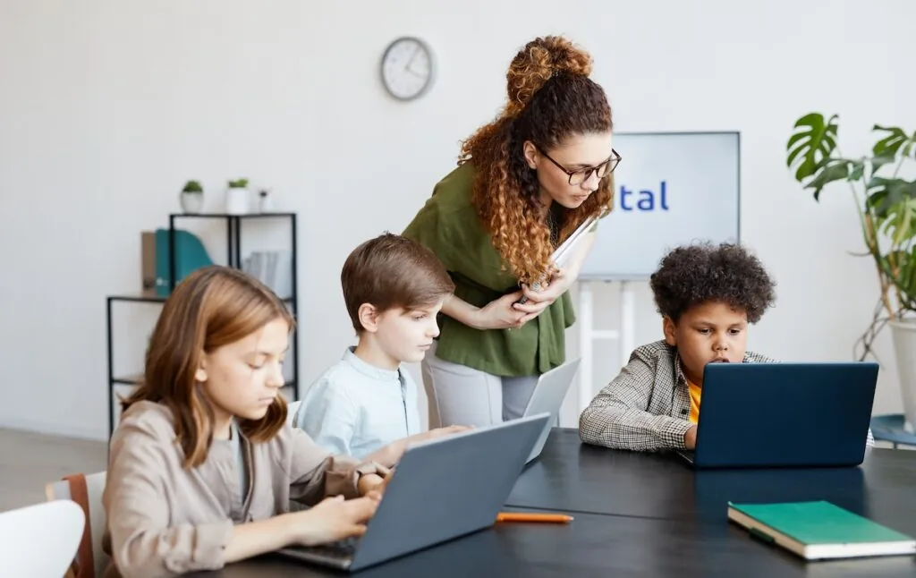 Three young students are sitting at a desk using their laptops, showcasing the concept of building equity in education. A female teacher stands behind and leans over one students computer to check their progress.