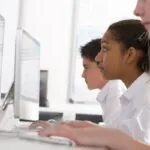 Side view of two female students and one male student sitting in a row in a computer class while looking at their desktop computers, showing the use of open educational resources in a school context.