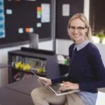 Une enseignante portant des lunettes, assise à un bureau, tenant un ordinateur portable et souriant, illustrant le concept de l'utilisation de l'IA pour faciliter la notation.