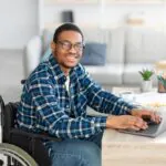 Smiling man wearing glasses sits in his wheelchair at a desk with his hands on a laptop while taking an exam designed for digital accessibility.
